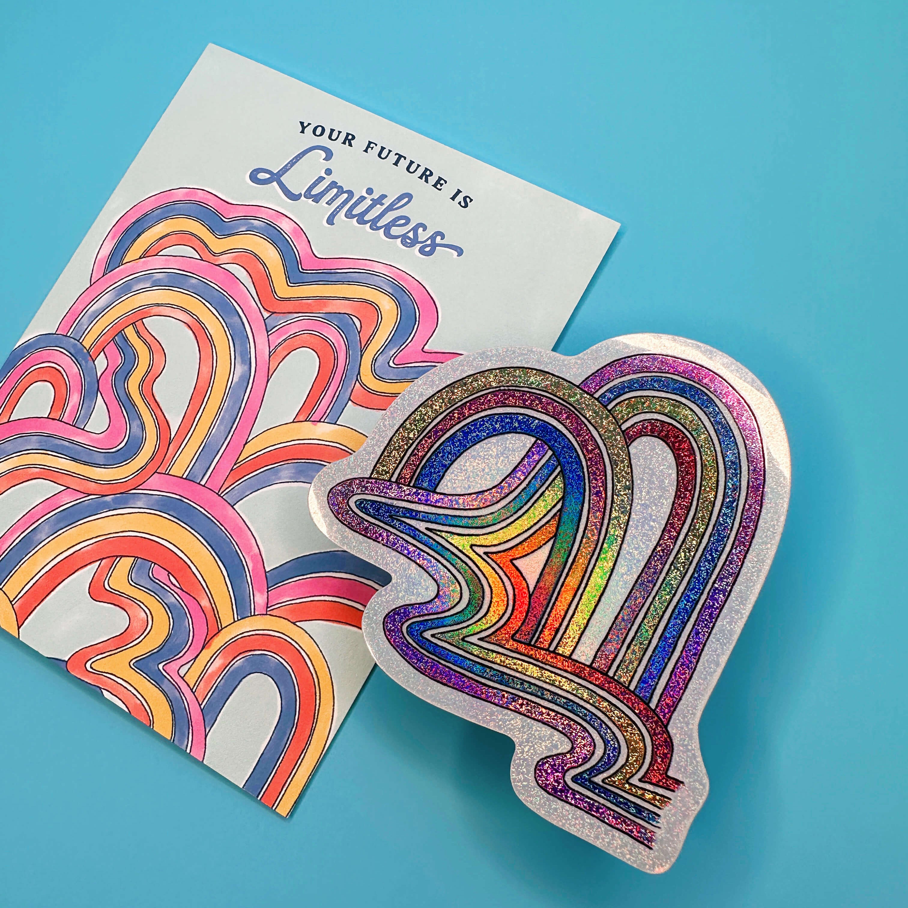 A glittery die-cut sticker shaped like a heart with rainbow-colored wavy lines, placed next to a white card printed with the same design and the words 'YOUR FUTURE IS Limitless' on a bright blue background.