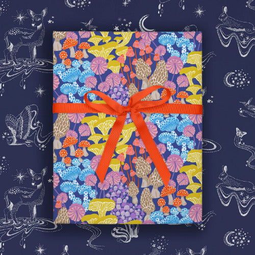 Gift box in a wrapping paper with a mushroom design tied with a red ribbon laying on top of a navy blue woodland animal celestial wrapping paper